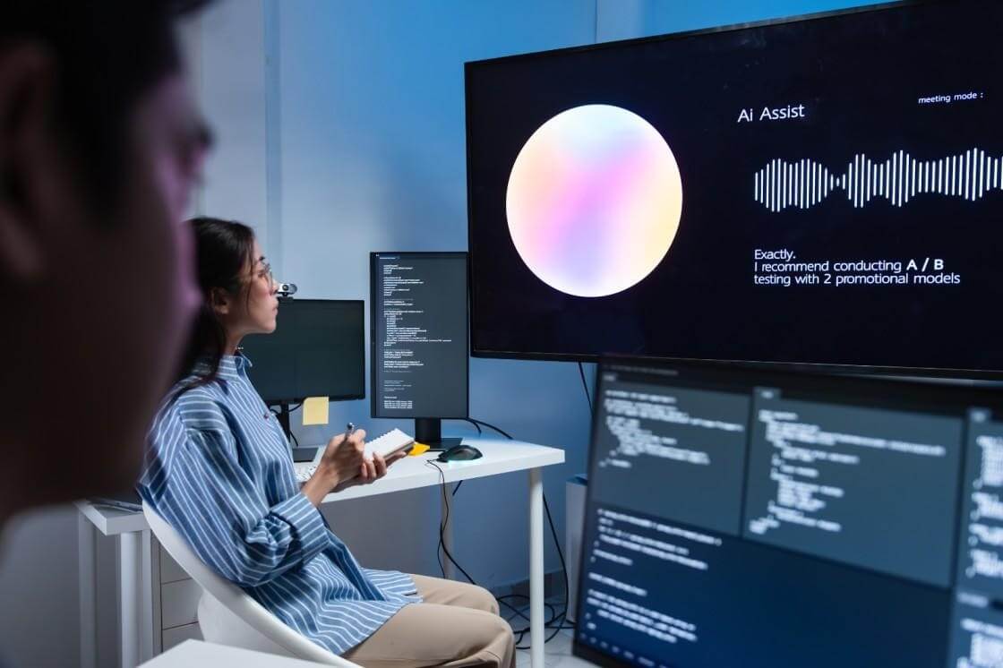 A woman sitting at a desk looking at multiple computer screens displaying code and an AI interface labeled Ai Assist with a waveform visualization