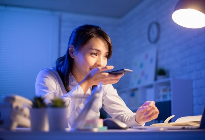 A woman sitting at a desk at night, speaking into her smartphone while looking at a computer screen