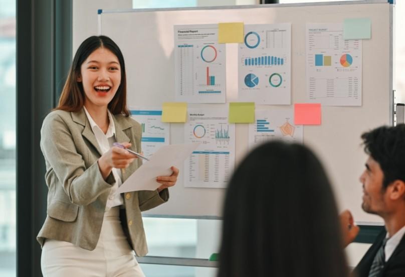 A woman in a business suit presenting in front of a whiteboard with charts and sticky notes, while two colleagues listen attentively